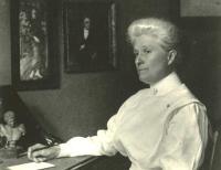 Anna Maxwell in white uniform sitting at a desk